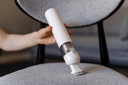 Woman cleans chair with cordless vacuum cleaner in living room space