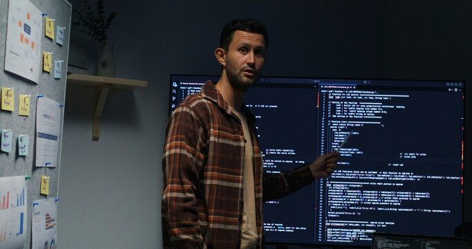 Man discusses software architecture and network solutions, reviewing code, data flows, cybersecurity and development roadmap in a corporate workspace.Nighttime Tech Strategy Session.