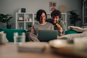 Happy couple enjoying online shopping with credit card and laptop at home, making secure internet...