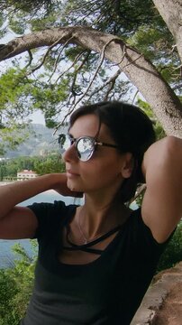 Portrait of pretty young tourist woman in sunglasses standing on top of mountain in front of amazing seascape enjoying vacation