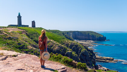 Woman traveler with backpack looking at Cap Frehel ( Cap d'Erquy) lighthouse and cliffs, Brittany,...