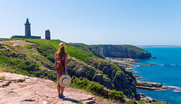 Woman traveler with backpack looking at Cap Frehel ( Cap d'Erquy) lighthouse and cliffs, Brittany, France