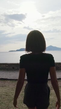 Back view of female traveler walking in the park and approaching the sea with open arms in front of amazing seascape at sunset, outdoors