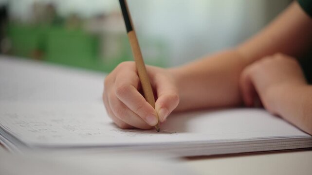 A child's hand carefully writes with a pencil in a lined notebook. The student is focused on their homework, practicing handwriting and learning.