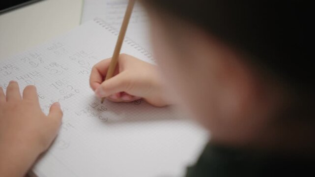 A child writes numbers with a pencil in a squared notebook while doing math homework. Over the shoulder view.