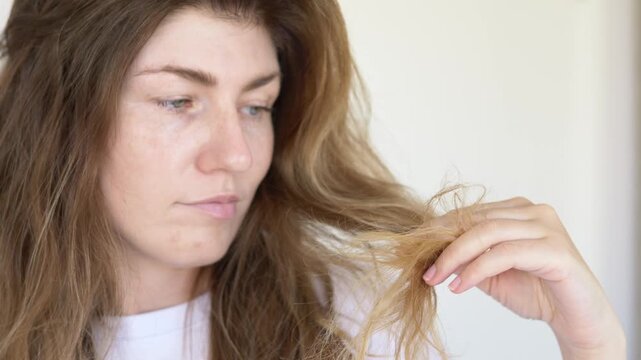 Close-up of female checking unhealthy hair strands with concerned expression. Hair damage, hydration treatment and keratin restoration concept for wellness, beauty salon and professional haircare