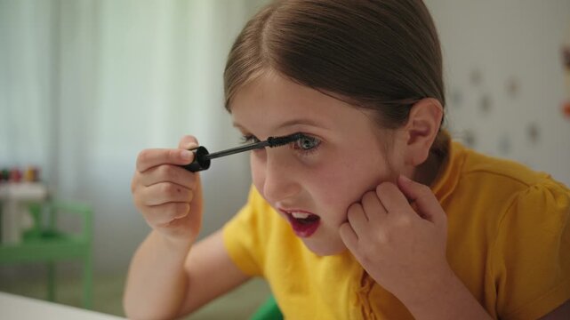A little girl with her mouth open in concentration carefully applies mascara to her eyelashes, experimenting with her mother's makeup for the first time.