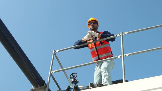 Male engineer with safety vest and helmet use walkie talkie to inspect rooftop cooling tower. Technician man checking building cooling system. A worker pointing while speaking into a walkie talkie.