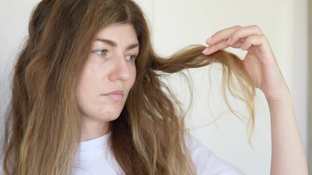 Woman looks at hair with worried concerned expression examining damage and loss in emotional haircare problem scene. Footage for trichology clinic promotion, hair loss treatment content, before-after