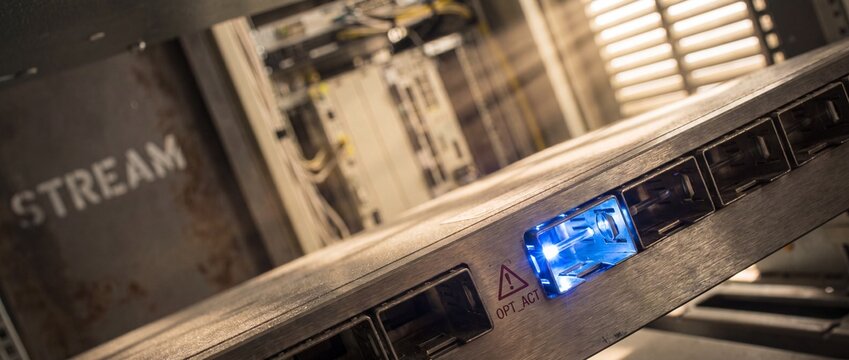 Data center network server switch with glowing blue optical port transmitting digital stream of information in modern hosting hub environment highlighting key cybersecurity and cloud database storage.