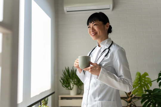 Smiling female doctor holding coffee mug by window in clinic. Healthcare professional, work-life balance, wellbeing and positive workplace concept