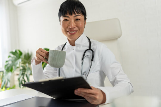 Smiling female doctor holding coffee mug while reviewing patient records in clinic. Healthcare administration, work-life balance and professional medical care concept