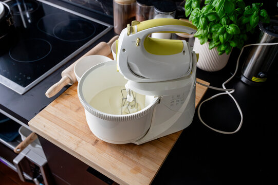 An electric hand mixer whipping fluffy white batter to soft peaks in a bowl on a wooden board in a modern marble kitchen preparing a homemade sponge cake.