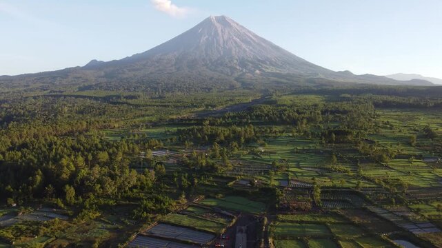 Drone view of Mount Semeru overlooking the agricultural landscape of Pronojiwo, East Java, with green farmland and volcanic scenery illuminated by morning light.