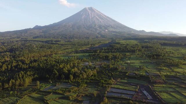 Drone view of Mount Semeru overlooking the agricultural landscape of Pronojiwo, East Java, with green farmland and volcanic scenery illuminated by morning light.