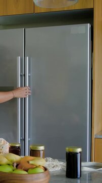 Vertical video: Picking up bottles, she in olive top opening steel fridge in kitchen, storing items