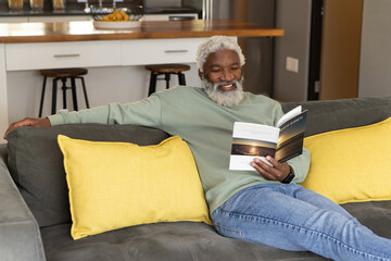 Obraz premium Senior African American man reclining on dark gray sofa at home reading paperback book