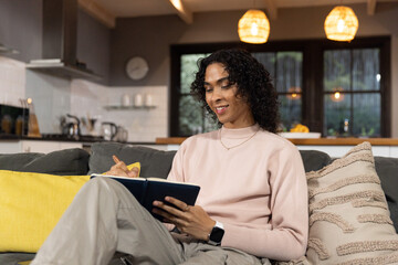 Non-binary person writing in dark-blue notebook and pen on gray sofa at home, wearing pink sweater © wavebreak3