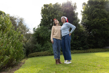Fototapeta premium Two female friends standing on grassy lawn wearing sweaters, jeans, Santa hat and antler headband