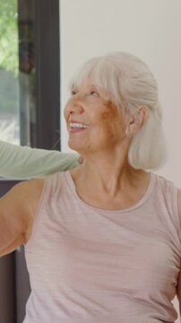 Vertical video: Smiling senior in pink top lifting arm on sofa as aide enters placing hand to aid