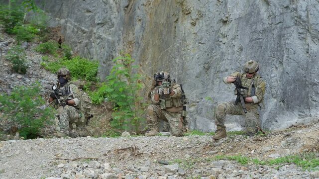Squad of five soldiers in camouflage uniform kneeling and aiming rifles during military tactical operation outdoor, special forces operators holding weapons near rock cliff