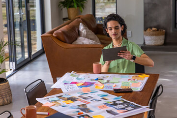 Obraz premium Asian man sitting at table at home holding tablet, reviewing layouts and sticky notes, copy space