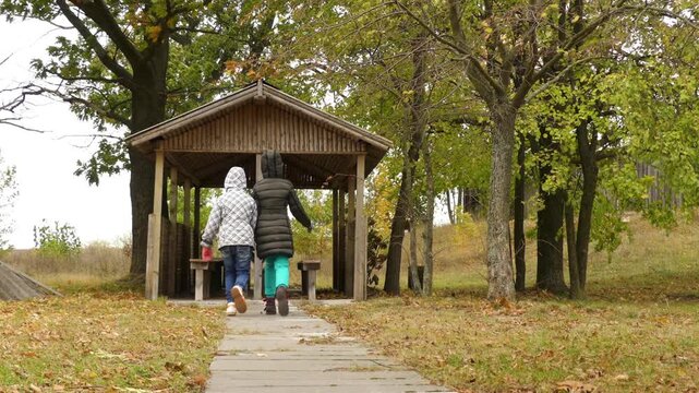 Two little girls sisters goes to wooden gazebo and sits down at his table in autumn leafy time.