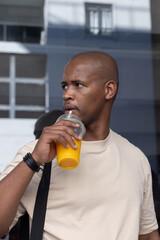 Fototapeta premium African man sipping juice from plastic dome-lid cup with straw, standing by glass facade