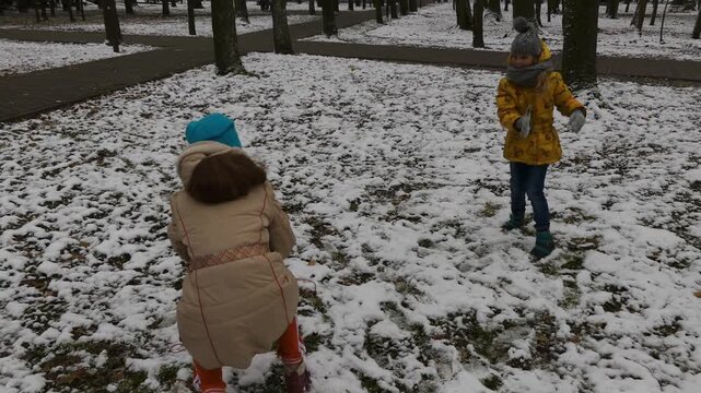 Two little sisters throw snowballs into each other in winter city park.
