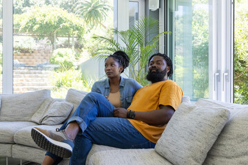 Obraz premium African couple sitting closely on light-gray sectional sofa near glass windows with potted palm