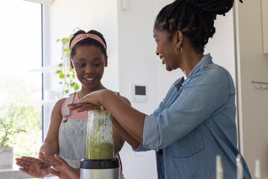 African American mom in denim pressing blender, teenage daughter with pink band gesturing by plants