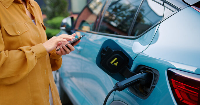 Close up of unrecognizable woman with a mobile phone in her hands uses a mobile app to connect a charging device to a depleted electric car at an outdoor charging station