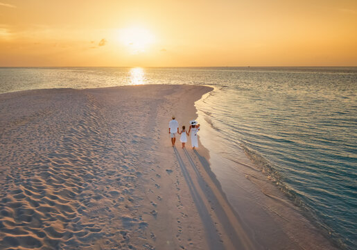 A beautiful holiday family in white summer clothing walks down a tropical paradise beach during sunset time