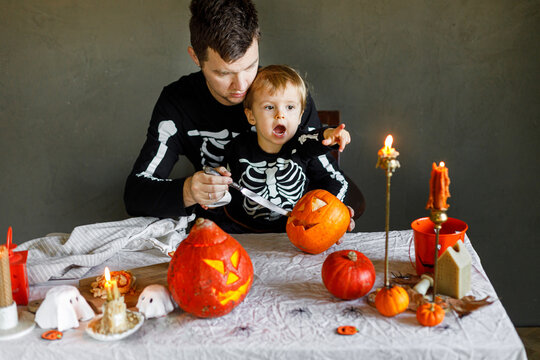 Happy Halloween. Father with child carving halloween pumpkin on table, making scary decorations on dark background. Space for text. Dad and son in skeleton costumes cutting jack o lantern together