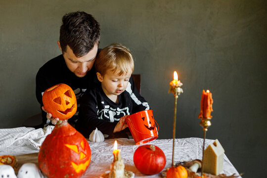 Dad and son in skeleton costumes making jack o lantern together. Happy Halloween. Father with child carving pumpkins on table with spooky decor and candles on dark background. Space for text