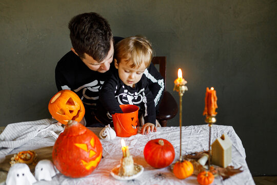 Dad and son in skeleton costumes making jack o lantern together. Happy Halloween. Father with child carving pumpkins on table with spooky decor and candles on dark background. Space for text