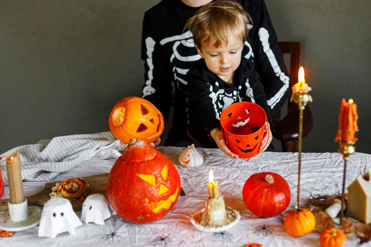 Dad and son in skeleton costumes making jack o lantern together and playing with bucket trick or treat. Happy Halloween. Father with child carving pumpkins on table with spooky decor and candles