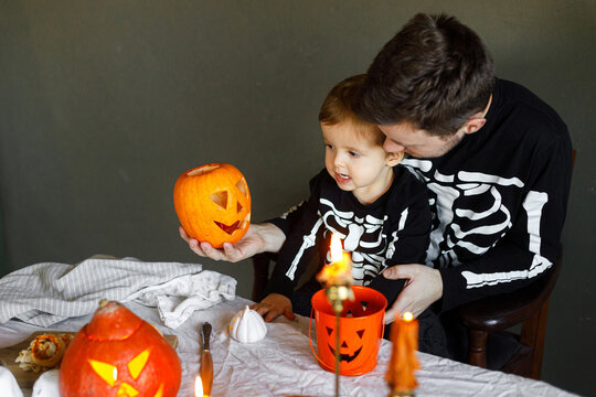 Happy Halloween. Father with child carving halloween pumpkin on table, making scary decorations on dark background. Space for text. Dad and son in skeleton costumes cutting jack o lantern together