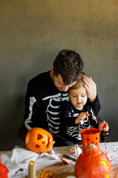 Dad and son in skeleton costumes making jack o lantern together and playing with bucket trick or treat. Happy Halloween. Father with child carving pumpkins on table with spooky decor and candles