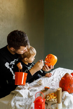 Happy Halloween. Father with child carving halloween pumpkin on table, making scary decorations on dark background. Space for text. Dad and son in skeleton costumes cutting jack o lantern together