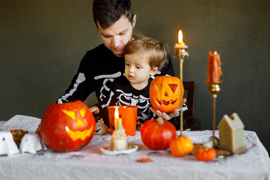 Dad and son in skeleton costumes making jack o lantern together. Happy Halloween. Father with child carving pumpkins on table with spooky decor and candles on dark background. Space for text