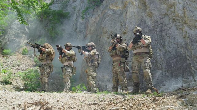 Squad of five soldiers in camouflage uniform standing and aiming rifles during military tactical operation outdoor, special forces operators holding weapons near rock cliff