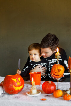 Dad and son in skeleton costumes making jack o lantern together. Happy Halloween. Father with child carving pumpkins on table with spooky decor and candles on dark background. Space for text