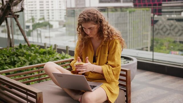 Curly-haired young woman sits on a balcony bench in a big city bakground, working remotely on her laptop . She pauses her work to answer a mobile phone call, smiling as she talks