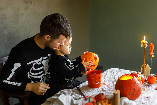 Happy Halloween. Father with child carving halloween pumpkin on table, making scary decorations on dark background. Space for text. Dad and son in skeleton costumes cutting jack o lantern together