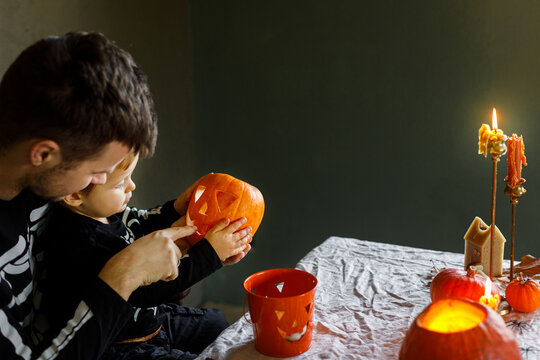 Dad and son in skeleton costumes making jack o lantern together. Happy Halloween. Father with child carving pumpkins on table with spooky decor and candles on dark background. Space for text