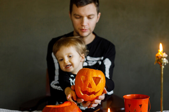 Happy Halloween. Father with child carving halloween pumpkin on table, making scary decorations on dark background. Space for text. Dad and son in skeleton costumes cutting jack o lantern together