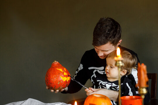 Dad and son in skeleton costumes making jack o lantern together. Happy Halloween. Father with child carving pumpkins on table with spooky decor and candles on dark background. Space for text