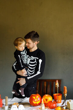 Dad and son in skeleton costumes standing at table with spooky decor and candles on dark background. Happy Halloween. Father with child carving pumpkins