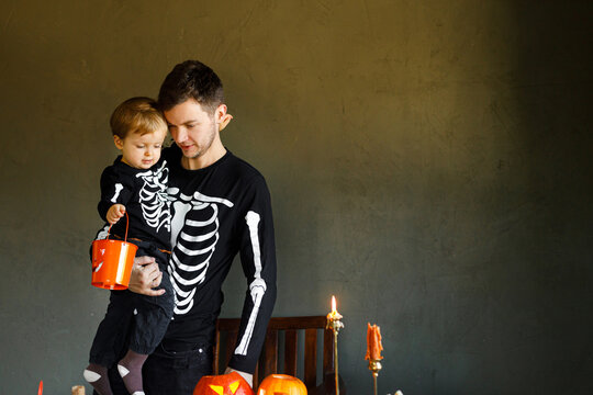 Happy Halloween. Father with child standing at carved halloween pumpkins and playing with bucket on dark background. Dad and son in skeleton costumes together trick or treat